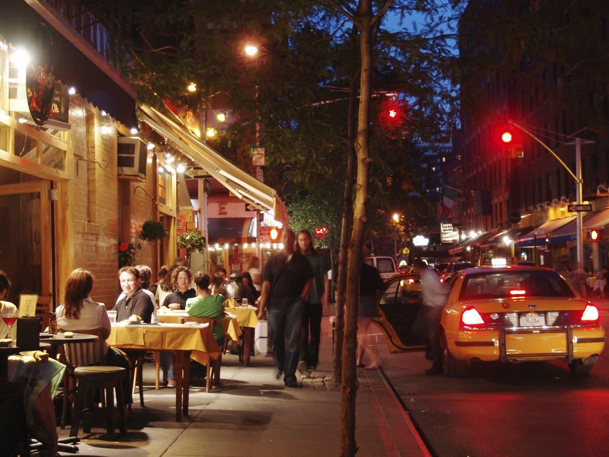Late-night NYC restaurant street scene with warm dining room lights, people walking after shows, concerts, games, and a night out