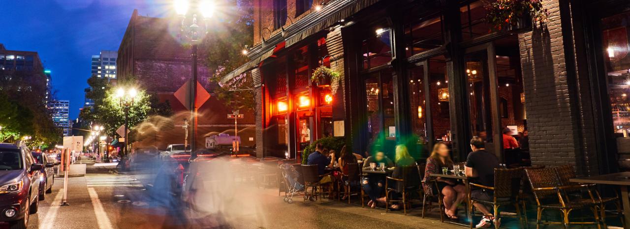 Late-night NYC restaurant street scene with warm dining room lights, people walking after shows, concerts, games, and a night out