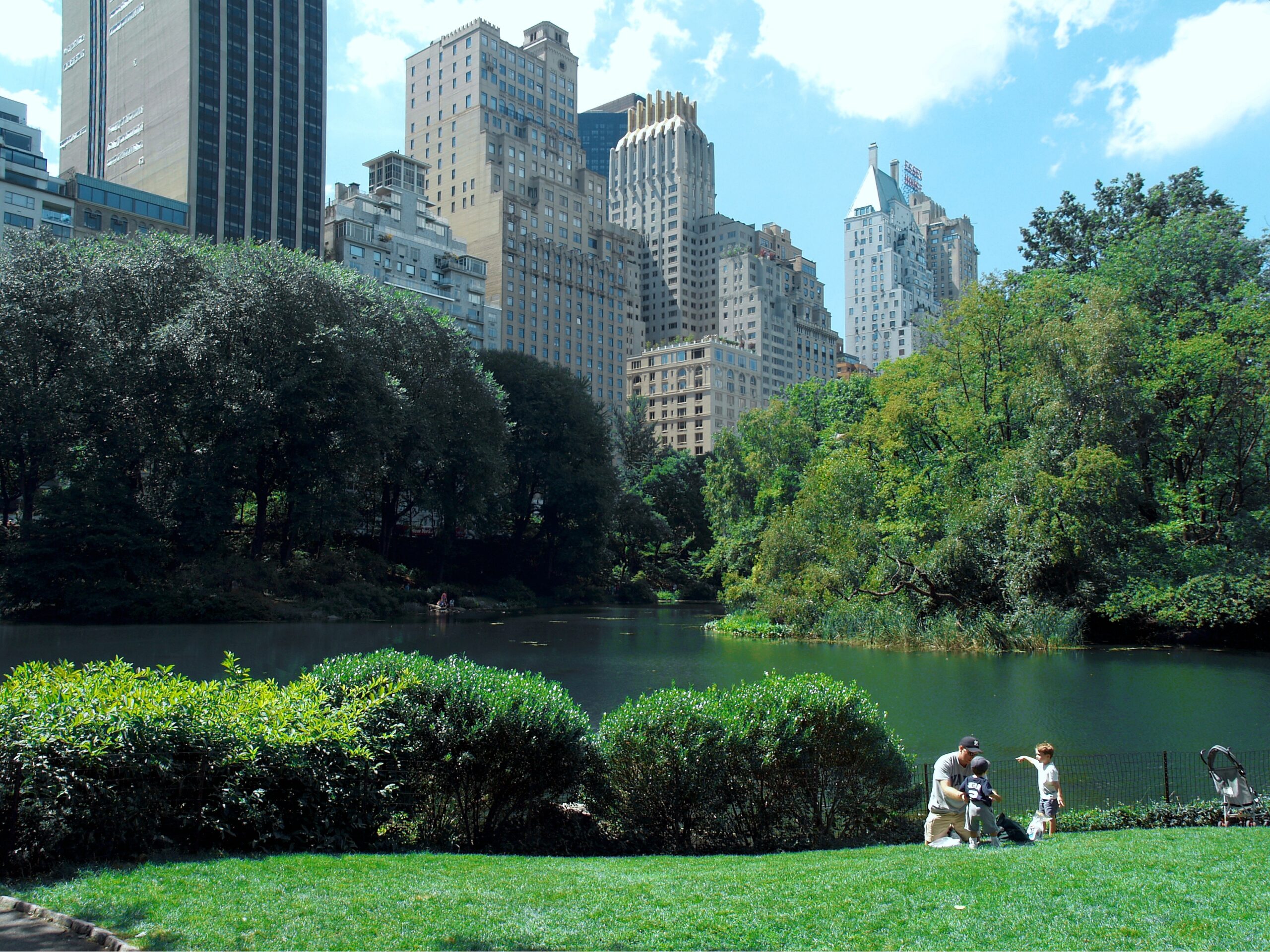 Family with children in Central Park with a stroller, pond, trees, and Midtown Manhattan buildings in New York City