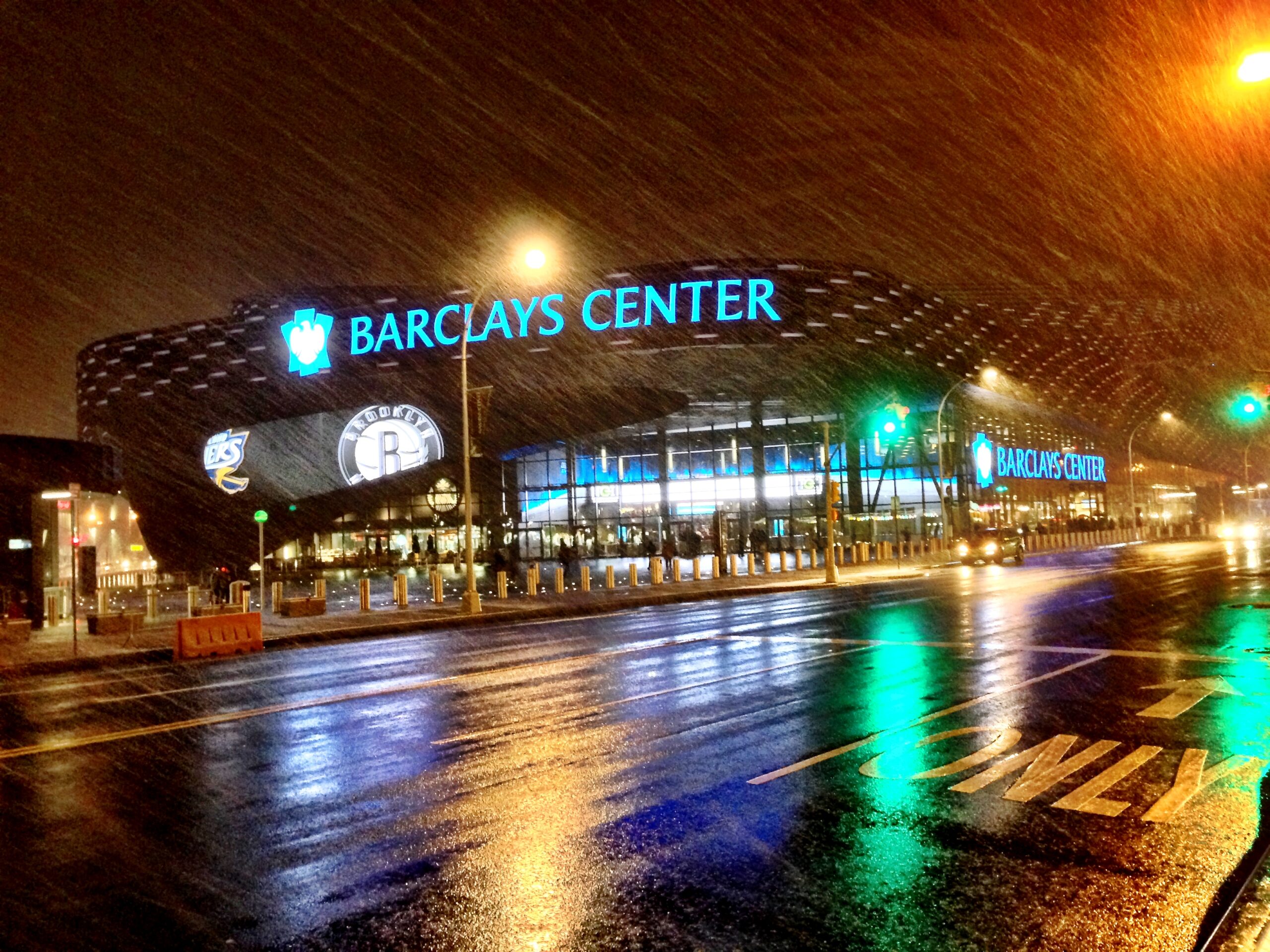 Barclays Center at night in Brooklyn with rain, bright arena signage, and wet street reflections before a concert, show, or game